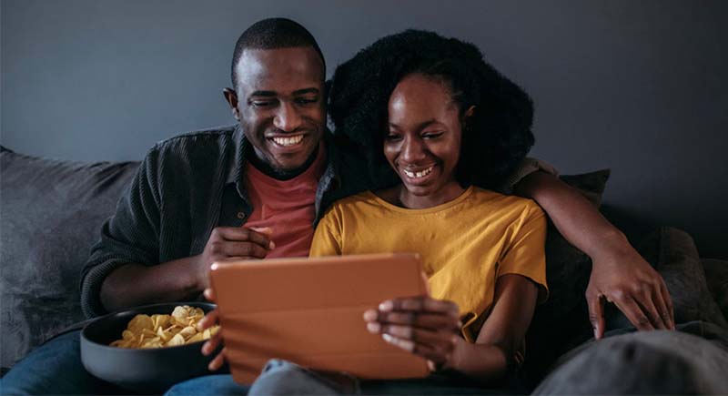 A young couple of color sits on a couch viewing a tablet